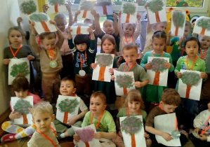 Dzieci z grupy pomarańczowej pozują do zdjęcia grupowego z otrzymanymi medalami i pracami plastycznymi wiosennego drzewa