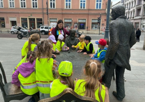 dzieci słuchają ciekawostek na temat pomnika Trzech Fabrykantów,