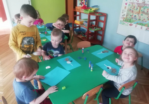 Dzieci siedzą przy stolikach, przyklejają elementy bociana do kartki. Starszy kolega pomaga dzieciom.