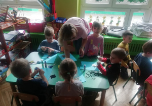 Dzieci siedzą przy stolikach, przyklejają elementy bociana do kartki. Pani Marta pomaga dzieciom.