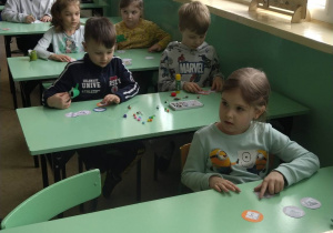 Dzieci siedzą w szkolnych ławkach i słuchają instrukcji nauczyciela