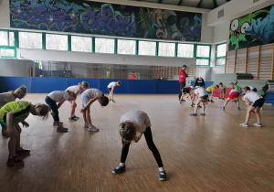 Dzieci wykonują skłony i ćwiczenia z piłką podczas zajęć sportowych w sali gimnastycznej