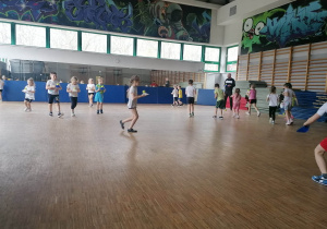 Dzieci biegają po sali gimnastycznej, trzymając piłkę umieszczoną w stożku, próbują utrzymać równowagę