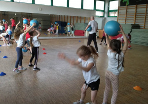 Dzieci ćwiczą w parach, stoją plecami do siebie i podają piłkę sposobem górnym i dolnym