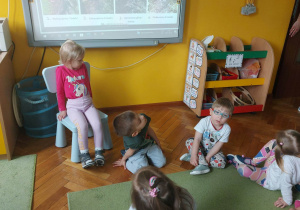 Dzieci siedzą na dywanie i oglądają prezentację na tablicy multimedialnej