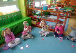 Dzieci siedzą na dywanie i uczestniczą w quizie, wykorzystując emblematy buziek