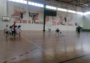 Dzieci ustawione są w rzędach, czekając na swoją kolej, dwójka dzieci wykonuje ćwiczenia ścigając się, biegną z piłkami