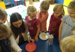 Dzieci z grupy żółtej stoją wokół stolika. Każdy ma słomkę i talerzyk wypełniony płynem, próbują robić bańki.