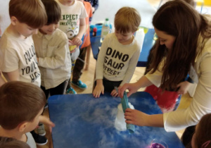Dzieci z grupy zielonej stoją wokół stolika, prowadząca przeprowadza eksperyment z suchym lodem.