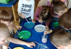 Dzieci z grupy zielonej stoją wokół stolika. Każdy ma słomkę i talerzyk wypełniony płynem, próbują robić bańki.