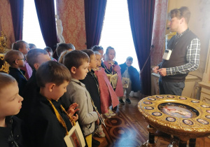 Dzieci przyglądają się eksponatom, przed dziećmi stoi przewodnik i opowiada ciekawostk