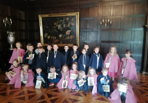 Grupa dzieci stoi ubrana w eleganckie starodawne stroje w sali balowej, dzieci trzymaj w rękach plakietki w wyszukiwanym skarbem