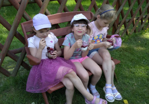 trzy dziewczynki siedzą na ławce w ogrodzie i jedzą podwieczorek