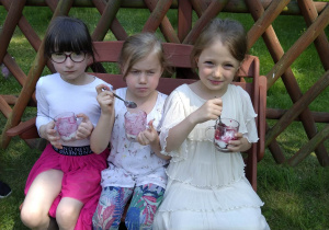 trzy dziewczynki siedzą na ławce w ogrodzie i jedzą podwieczorek