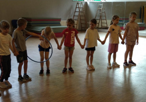 dzieci stoją w szeregu, trzymając się za ręce starają się przekładać hula-hop