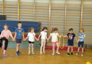 dzieci stoją w szeregu, trzymając się za ręce starają się przekładać hula-hop