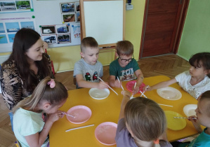 Dzieci zamaczają słomkę w płynie do baniek i dmuchając na talerzyk robią bańki.