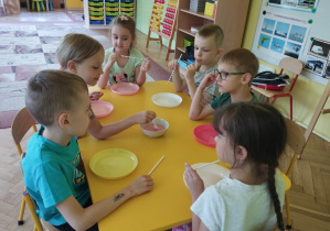 Dzieci siedzą przy stolikach, przed sobą mają plastikowe talerzyki, w rękach trzymają słomki. Na środku stoi miska wypełniona woda z płynem do mycia naczyń, jedna osoba miesza płyn do baniek.