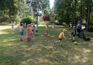 Grupa dzieci stoi w rzędzie i wykonuje ćwiczenia z piłkami