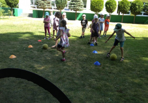 Grupa dzieci stoi w rzędzie i wykonuje ćwiczenia z piłkami