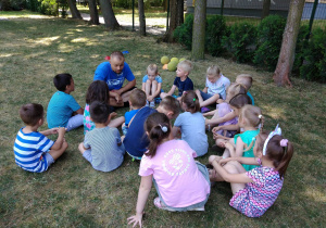 Dzieci wraz z trenerem siedzą na trawie i omawiają zasady zabawy