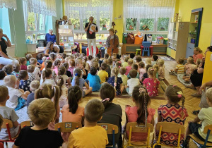 Dzieci z całego przedszkola siedzą na krzesłach i na dywanie i słuchają koncertu