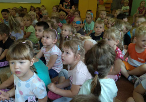Dzieci z całego przedszkola siedzą na krzesłach i na dywanie i słuchają koncertu
