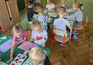 Dzieci siedzą przy stolikach i malują farbami za pomocą palców u dłoni pracę plastyczną