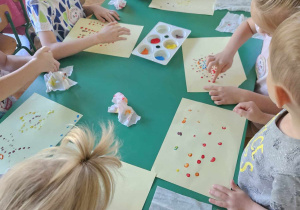 Dzieci siedzą przy stolikach i malują farbami za pomocą palców u dłoni pracę plastyczną