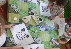 Dzieci siedzą przy stolikach i malują farbami kota za pomocą palców u dłoni
