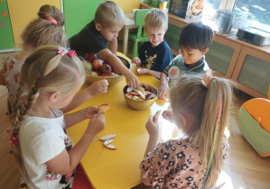 Dzieci siedzą przy stoliku i nawlekają na igłę kawałki jabłek.