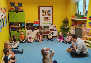 Dzieci siedzą na dywanie, odpowiadają na zagadki zadane przez aktora.