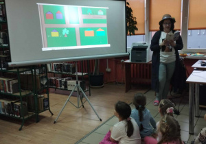 Prowadząca przebrana za Mary Poppins odczytuje dzieciom fragment książki, w tle ilustracja. Dzieci siedzą na poduszkach.