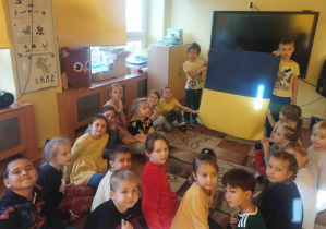 dzieci siedzą na dywanie, dwóch chłopców i dwie dziewczynki trzymają flagę Ukrainy. Dzieci opowiadają ciekawostki, które zapamiętały podczas prezentacji multimedialnej o Ukrainie.