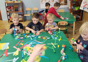 Dzieci siedzą przy stole i kleją choinkowy łańcuch z papierowych kolorowych pasków