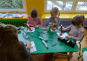 Dzieci podzielone na grupy siedzą przy stolikach i wykonują wzory na szablonach przy użyciu długopisów 3D.