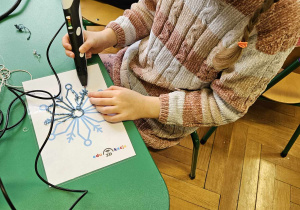 Dzieci podzielone na grupy siedzą przy stolikach i wykonują wzory na szablonach przy użyciu długopisów 3D.
