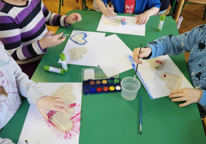 Dzieci siedzą przy stolikach, wykonują pracę plastyczną przy użyciu serca z gazety oraz farb akwarelowych.