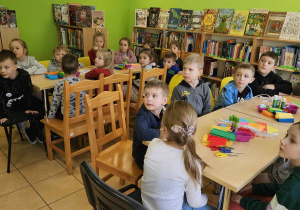 Dzieci siedzą przy stolikach i słuchają książki.