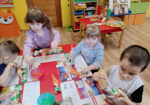 Dzieci siedzą przy stoliku, malują farbami marchewki wykonane z papieru