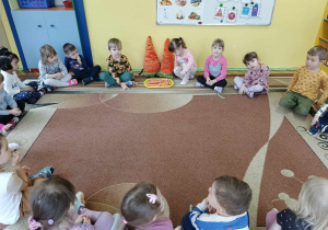 Dzieci siedzą w kole na dywanie, obok nich marchewka karotka wraz z siostrą