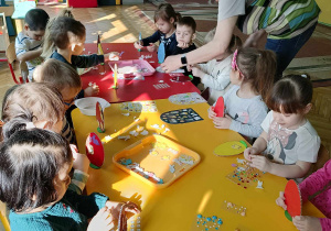 Dzieci siedzą przy stole, przyklejają gotowe elementy do jajka