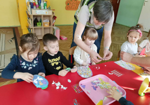 Dzieci siedzą przy stole, przyklejają gotowe elementy do jajka