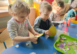 Dzieci siedzą przy stole, przyklejają gotowe elementy do jajka