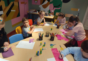 Dzieci siedzą przy stolikach, projektują stroje wykorzystując przy tym, skrawki wzorzystych papierów, bibuły i innych przyborów plastycznych.