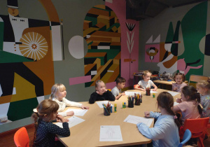Dzieci siedzą przy stolikach, projektują stroje wykorzystując przy tym, skrawki wzorzystych papierów, bibuły i innych przyborów plastycznych.