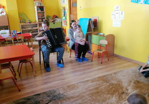 Chłopiec gra na akordeonie, obok niego na krzesełku siedzi kobieta z dzieckiem na kolanach, dzieci słuchaj muzyki siedząc na dywanie