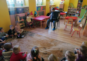 Dzieci siedzą na dywanie tworząc półkole, jedna dziewczynka siedzi na krześle, chłopiec gra na akordeonie