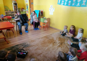 Chłopiec gra na akordeonie, obok niego na krzesełku siedzi kobieta z dzieckiem na kolanach, dzieci słuchaj muzyki siedząc na dywanie