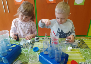 Dzieci siedzą przy stoliku i barwią wodę kolorowym tuszem.